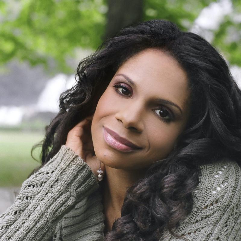 Singer Audra McDonald, wearing a grey sweater, rests her head on her hand on a stone fence outdoors.