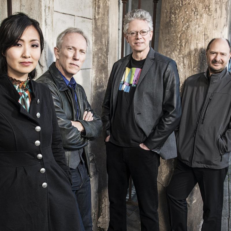 The musicians of the performing group Kronos Quartet in front of a grey stone wall and iron railing.