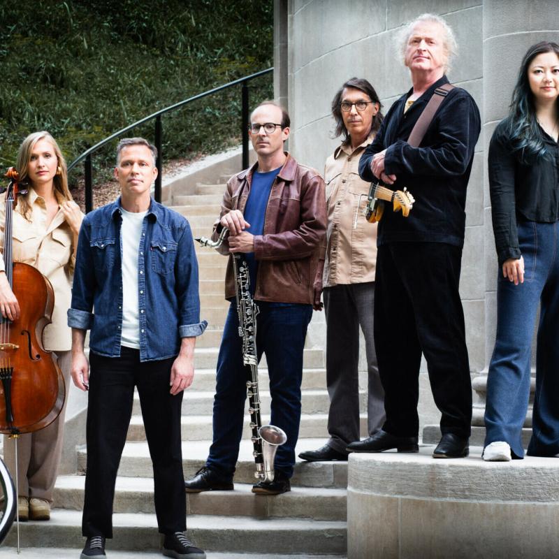 The six performers of Bang on a Can All Stars stand across a set of steps with a railing running down the left side. The Cellist and the Bass Clarinetist have their instruments. 