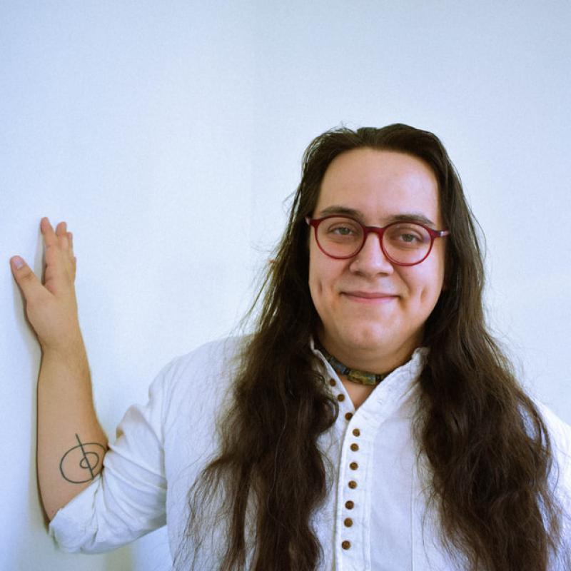 Composer inti giffis-visueta stands with their hand against a white wall wearing a long-sleeved white button shirt.