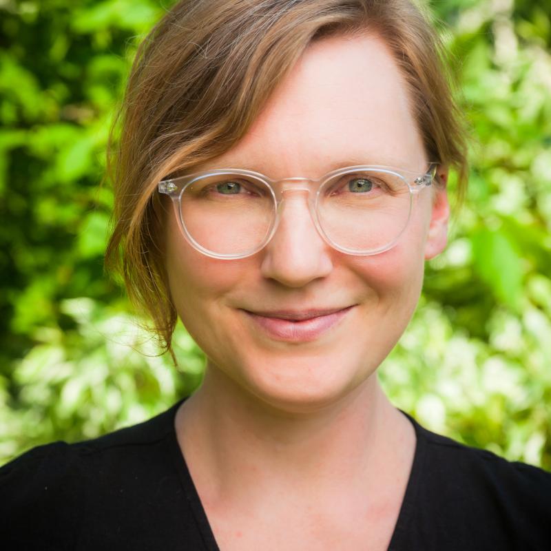Anna Reiser smiles, wearing glasses and a black shirt, with green foliage in the background on a sunny day. 