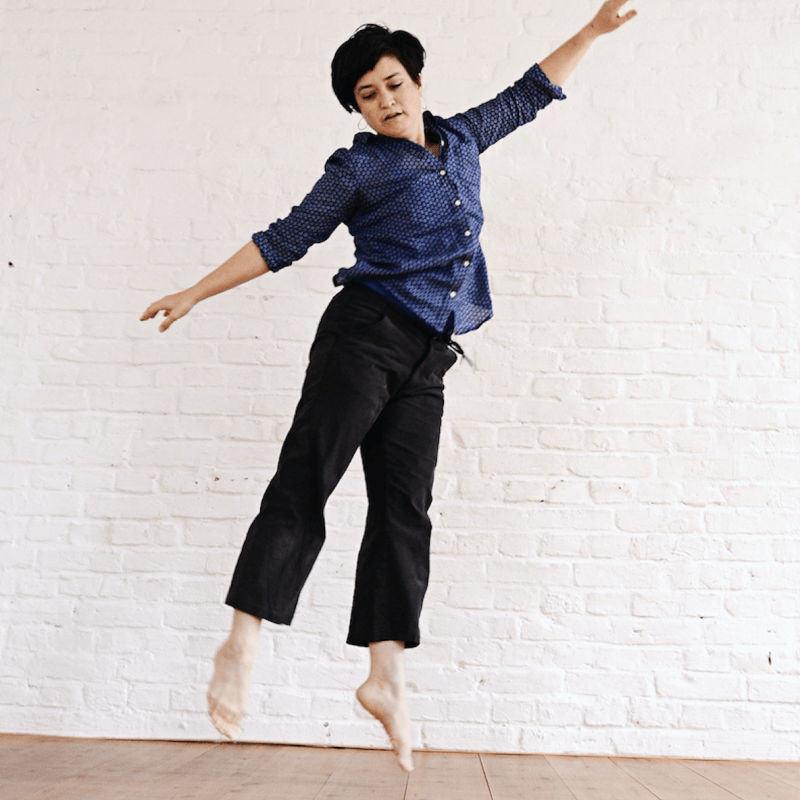 full body image of black haired woman caught mid-jump, with legs and arms outstretched, slightly curved and spiraled spine, looking down. photo by Manuel Miethe