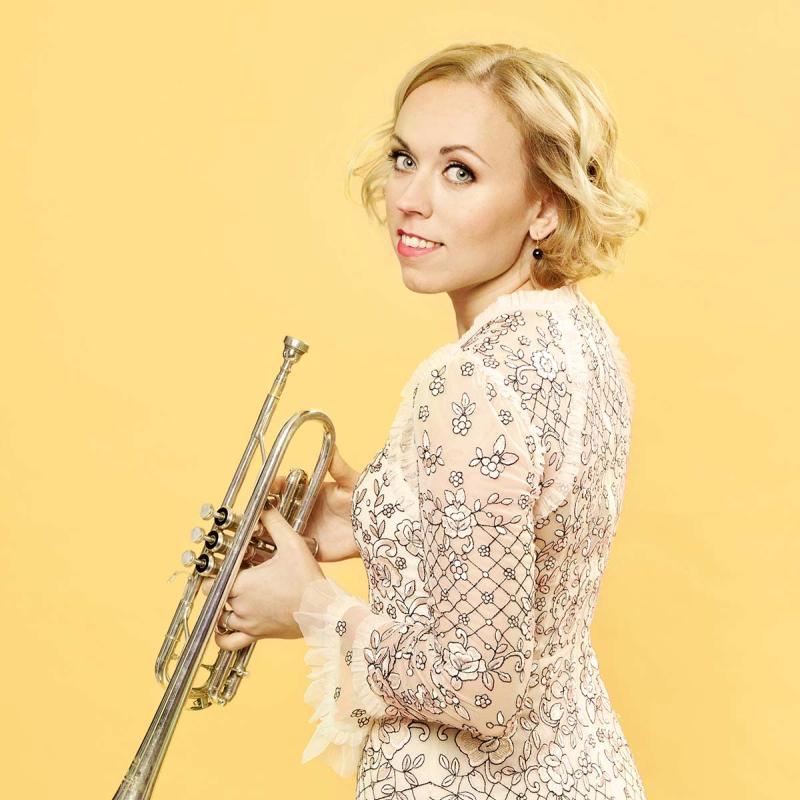 Trumpet player Tine Thing Helseth holds her trumpet looking back at the camera over her left shoulder against a yellow background. 