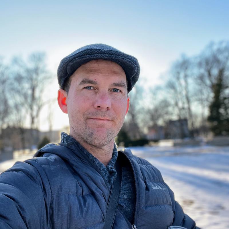 Picture of man with cap, facing camera in Appleton City Park, with winter sun casting long afternoon shadows in the background