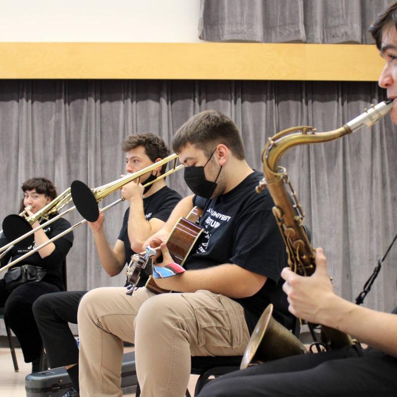 Four students with various instruments, such as saxophone and trumpet, sit in a row in a large room.