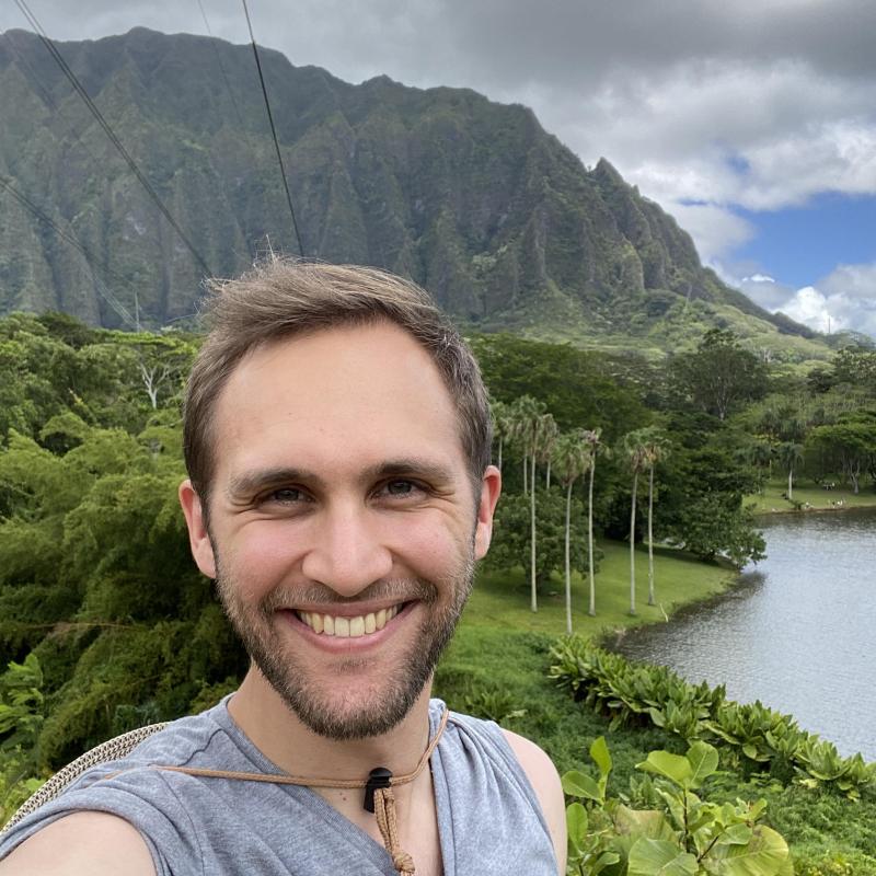 Joe Johnson '17 in a casual grey shirt, smiling in front of a lake, mountain, and greenery at Ho'omaluhia Botanical Gardens on O'ahu