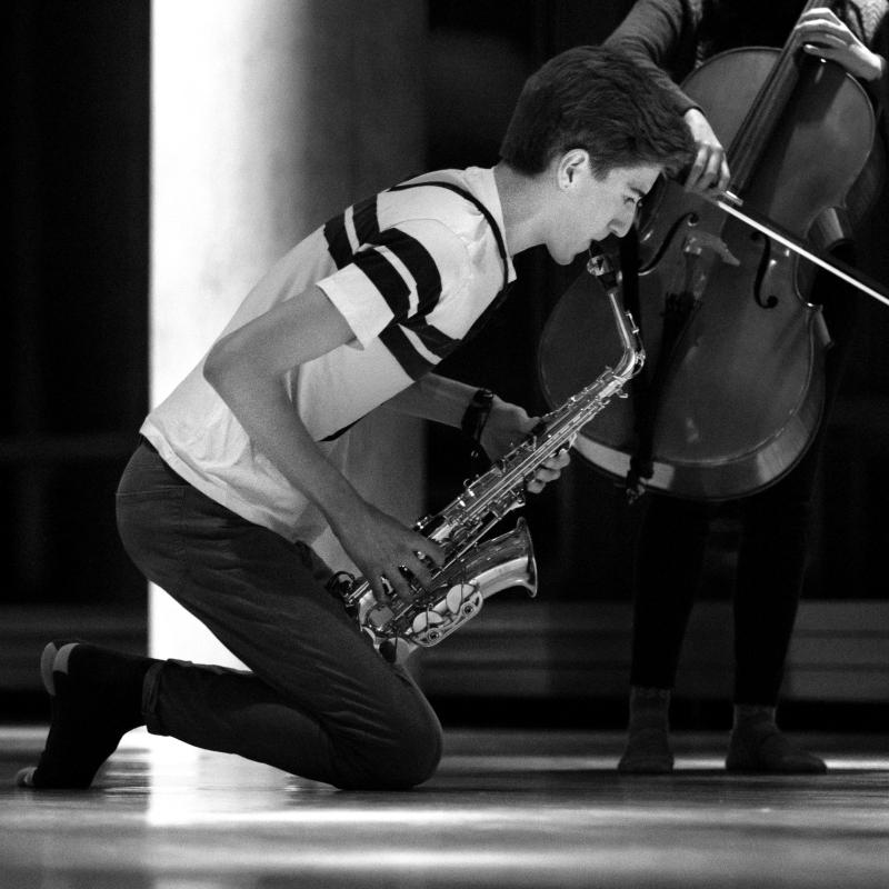 Boy plays saxaphone kneeling on floor in front of cello player.