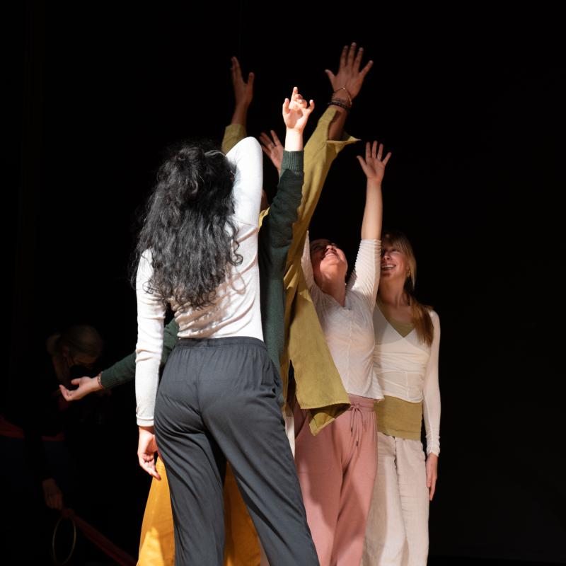 Dancers stand in a clump with arms reaching upwards in a dark room.