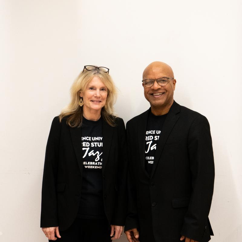 Black man and white women stand next to eachother on white wall wearing black outfits with t-shirts with white writing.
