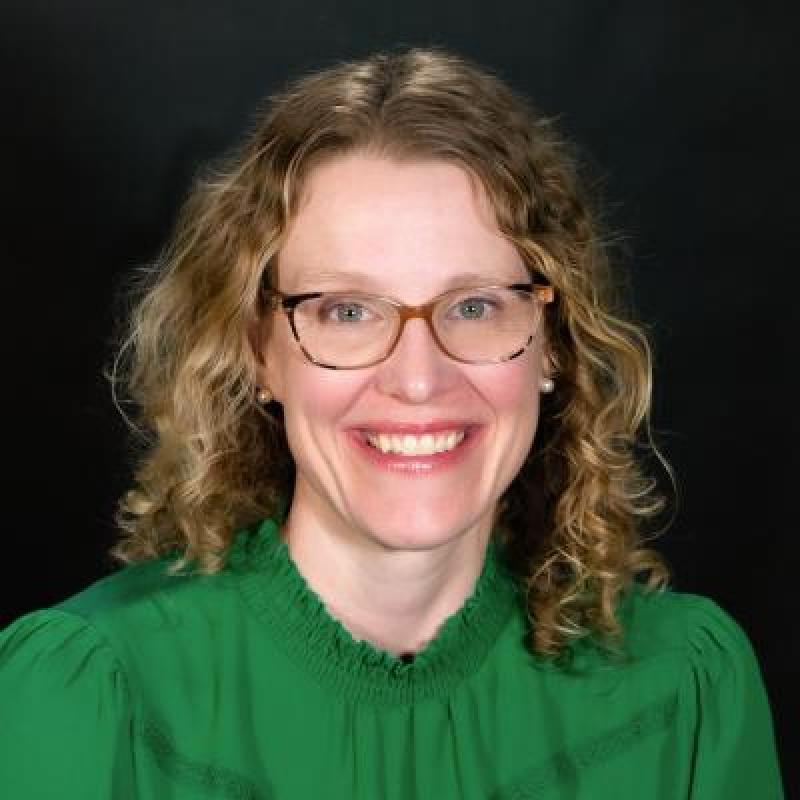 Betsy Schlabach pictured with glasses and blonde hair. Sitting and facing camera. 