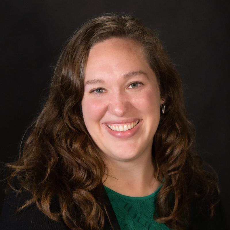 a teacher with long brown hair wearing a green shirt and black blazer smiles at the camera