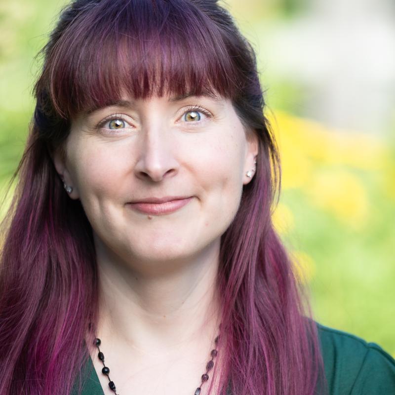 Angela outside, portrait by Rachel Crowl. Angela has green eyes, white skin tone, long magenta hair, and is smiling.