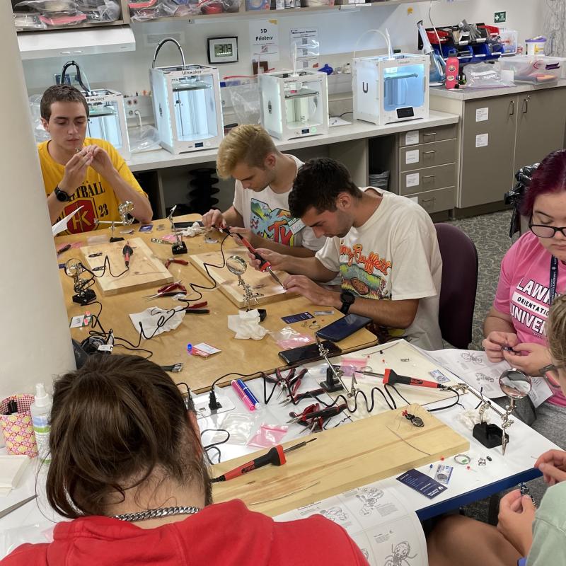 Students sitting around a table practicing soldering in the makerspace with 3D printers printing in the background.