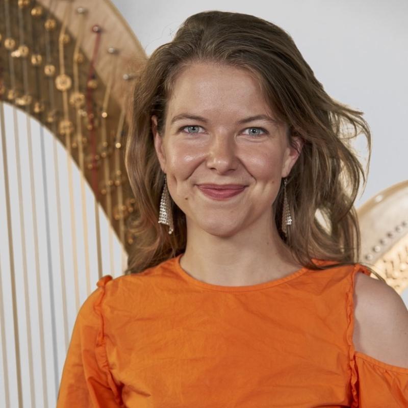 Johanna smiles at the camera, she has long hair and is wearing an orange shirt. She stands in front of a full sized pedal harp.