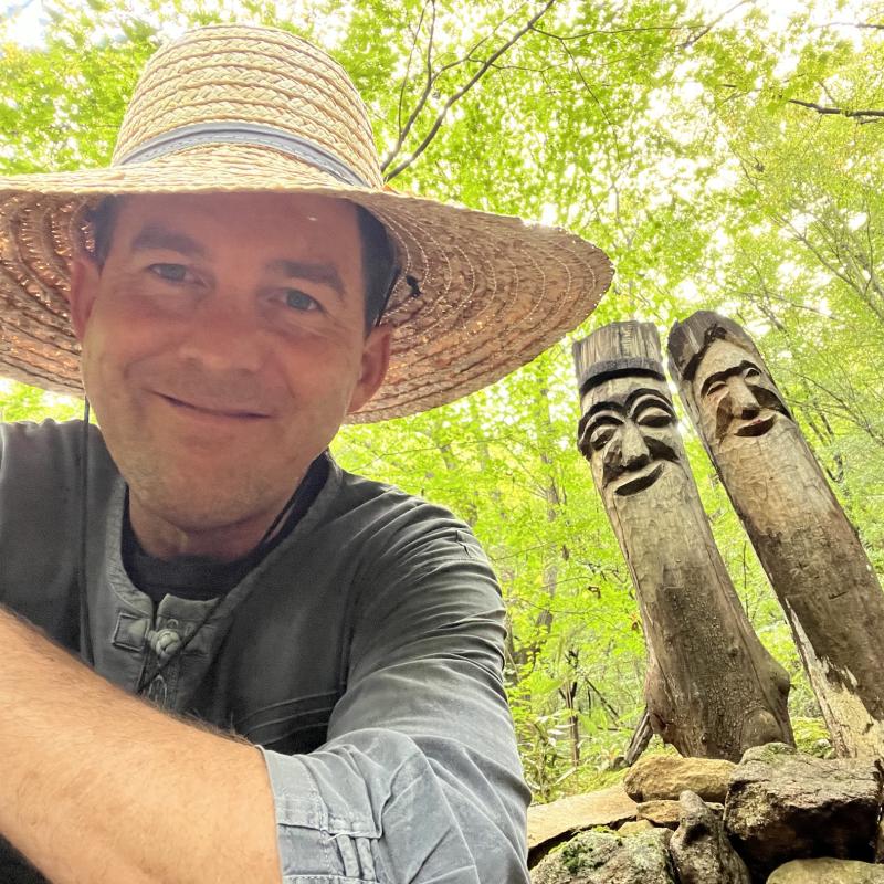 a man in a hat smiles in front of two wooden totems behind his shoulder on a mountain side