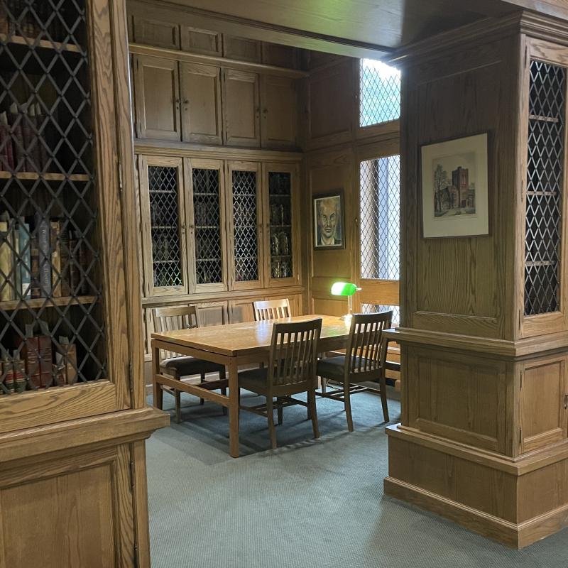 Wooden table in a with a green lamp on top, surrounded by wooden cabinets and bookshelves with metal lattice doors.