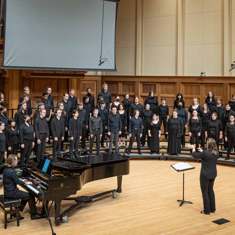 A choir dressed in concert black accompanied by a pianist sings on risers in the chapel while their director gestures in front of the group