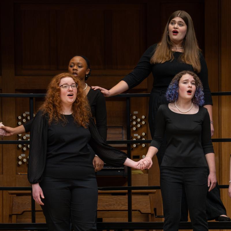 Four women in concert black sing on stage, two holding hands and one touching her neighbor's shoulder