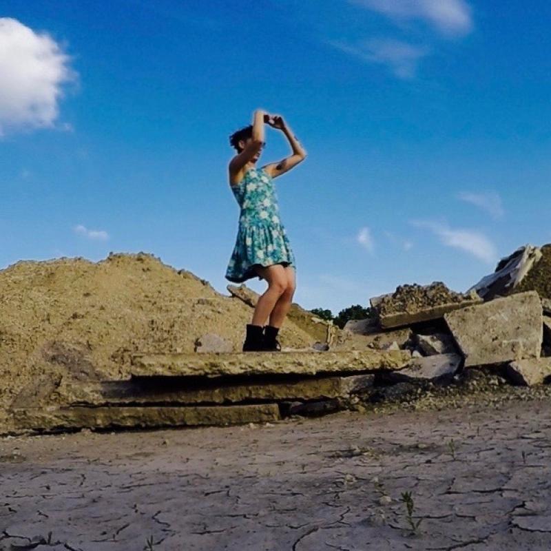 mauriah stands atop a pile of rock, bright blue sky and cracked earth surrounds. she wears a blue dress that stops above the knee, black boots. her hands curl like binoculars over her eyes. she looks out towards horizon.