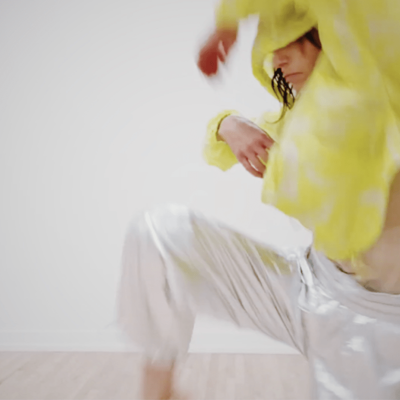 mauriah is caught mid action. she is lifting her right leg up, knee bent while curling arms inward, right arm into chest, left arm up overhead. Mauriah is wearing baggy silver pants, a bright yellow windbreaker, hood up. The hood obscures her face, only nose and mouth are visible. A shock of dark brown hair falls over face.