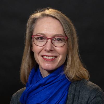 A head shot of a middle-aged, smiling white woman with red glass, wearing a blue scarf and gray sweater