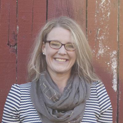 (white woman with blonde hair and dark rimmed glasses wearing a green scarf and striped navy blue top standing infant of a red wooden wall)