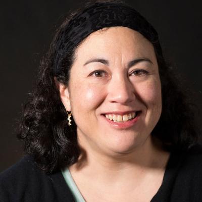 the head and shoulders of a smiling, dark haired woman against a black background. She is wearing a light green top and a black cardigan sweater.