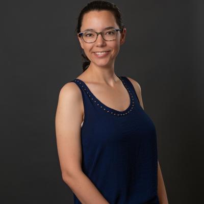 Middle-aged white woman with blue tortoise shell glasses and dark brunette hair pulled back in a ponytail. She is wearing a royal blue, sleeveless blouse