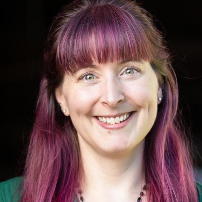 Angela with a black background, portrait by Rachel Crowl. Angela has green eyes, white skin tone, long magenta hair, and is smiling.