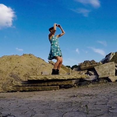mauriah stands atop a pile of rock, bright blue sky and cracked earth surrounds. she wears a blue dress that stops above the knee, black boots. her hands curl like binoculars over her eyes. she looks out towards horizon.