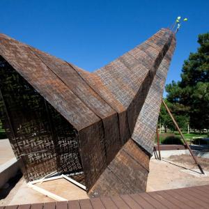 Giant sculpture with perforated and patinaed steel panels and geometric beadwork designs on them, overlap like a crane's feathers.
