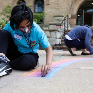 Student residence hall adviser sitting and crouching on ground to draw rainbow.