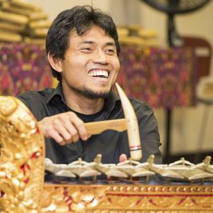 Smiling Balinese man with a short goatee playing a metallophone with a wooden mallet.