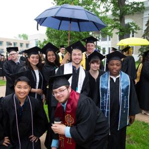 A group of Lawrentians in gap and gown smile while more graduates stand chatting with each other in background.