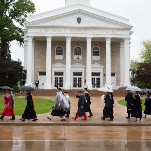 Lawrentians with umbrellas and wearing clear plastic ponchos, walk in two lines down College Ave. with Memorial Chapel in the background.