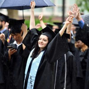 A graduate stands, smiling, with both hands raised.