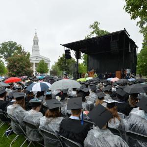 Graudates sit with ponchos and umbrellas in front of stage on Main Hall Green during Commencement.