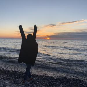 Silhouette of person with arms raised against a setting sun along a lakeshore