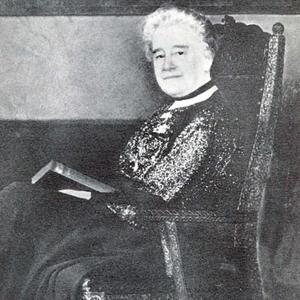 black and white photo of an older white woman in victorian style dress sitting in an ornate chair with an open book. 