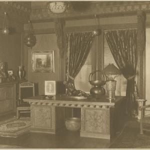 a sepia toned photograph of an ornate interior with a desk, art objects, heavy curtains, and lamps.