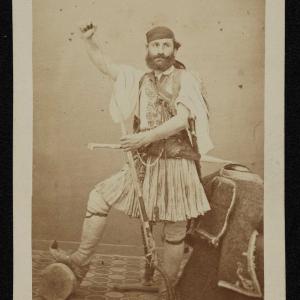 black and white studio photo of a white man wearing clothes from another culture, with an arm raised and a knife in his hand.