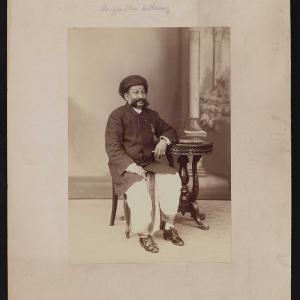a black and white studio photo of an Indian man in a turban and kurta seated at a carved wooden side table.