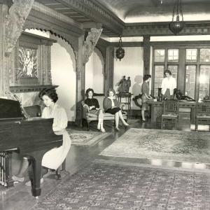 black and white photo of young white women playing piano and relaxing in a room with ornate carvings and rugs.