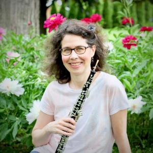 A smiling bespectacled woman holding an oboe, set against some flower bushes.