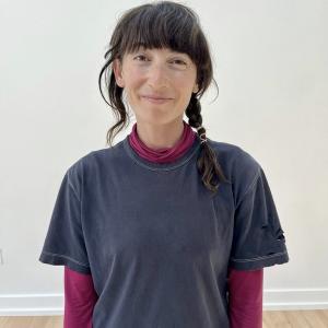 mauriah faces forward arms by her side. she is smiling, mouth closed. her skin is light, brown hair covers forhead and a braid comes around her left shoulder. she is wearing a baggy blue tshirt and a magenta turtle neck underneath. behind her, there is a grey white wall, light hardwood floors.