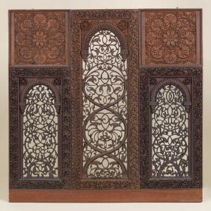 Decorative carved wooden screen with three vertical panels. The center panel features an arched opening filled with intricate scrollwork, while the two side panels contain matching carved lattice designs beneath floral-patterned panels. The entire piece is covered with detailed vine and floral carvings.