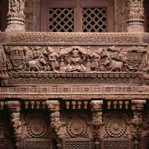 Intricately carved wooden balcony or façade with a central seated figure surrounded by smaller figures, animals, and decorative patterns. The structure is covered in detailed floral, geometric, and architectural carvings.