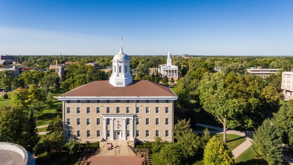 Aerial view of a campus building with a white cupola, surrounded by trees and other academic buildings under a clear blue sky.