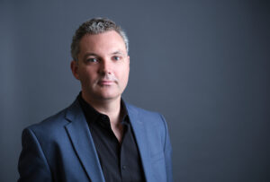 A man with short, curly hair wears a blue blazer and black shirt, standing in front of a plain gray background.