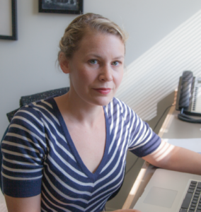 A person in a striped shirt sits at a desk with a laptop, looking toward the camera. Sunlight and shadows are visible on the wall and desk.